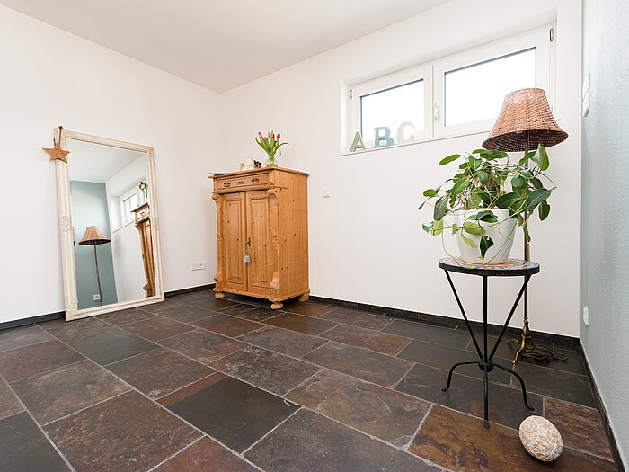 Heller Raum mit dunklem Steinboden, Holzschrank, Pflanze auf Beistelltisch, Spiegel an der Wand.