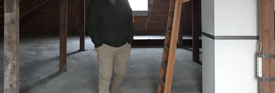 Mann in unfertigem Rohbau mit Holzstützen und Leiter im Bauinneren.