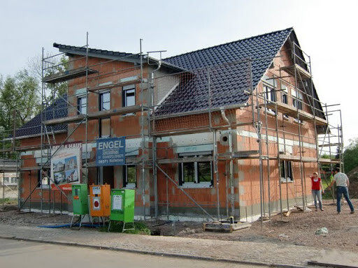 Zweistöckiges Haus im Rohbau mit Gerüst, Ziegelwänden und Dach; Mülltonnen im Vordergrund.