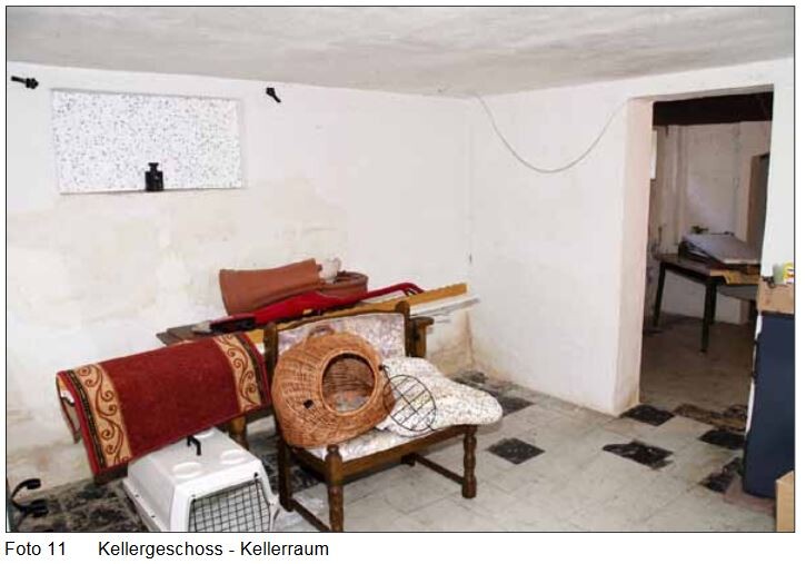 Kellerraum mit weißen Wänden, Möbeln, Teppichrollen, Körben und einem Stuhl; Durchgang zu einem weiteren Raum.