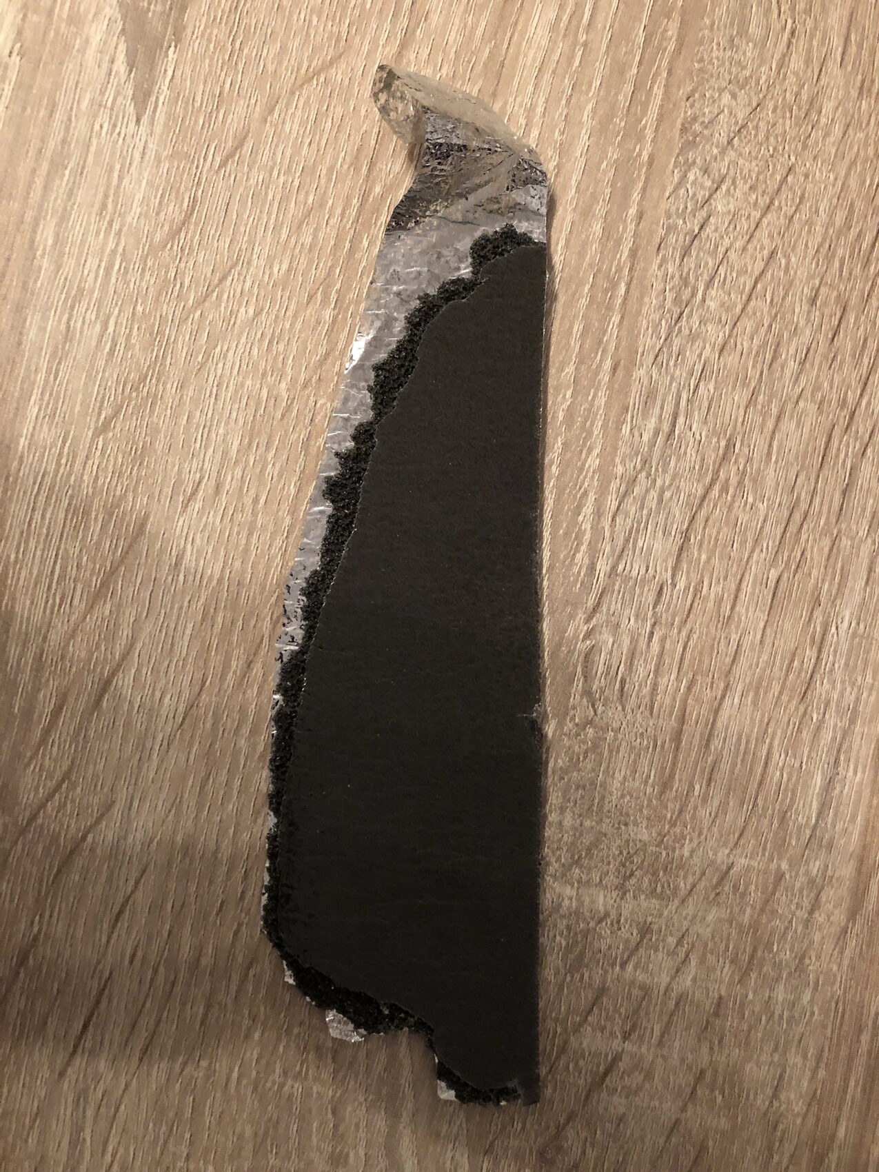 Fragment einer schwarzen Bitumen-Dachbahn mit rauer Kante auf Holzuntergrund.