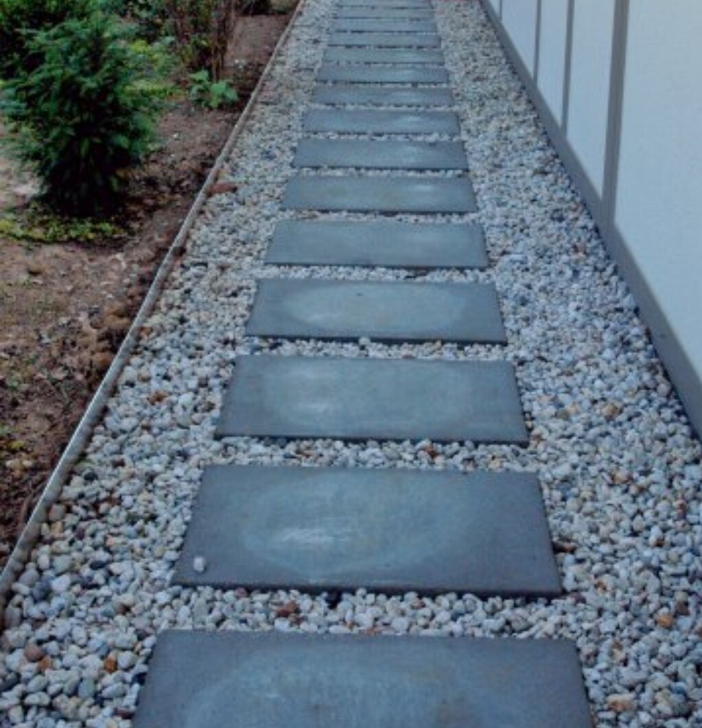 Gartenweg aus quadratischen Steinplatten auf Kies, neben einer Hauswand, links Sträucher.