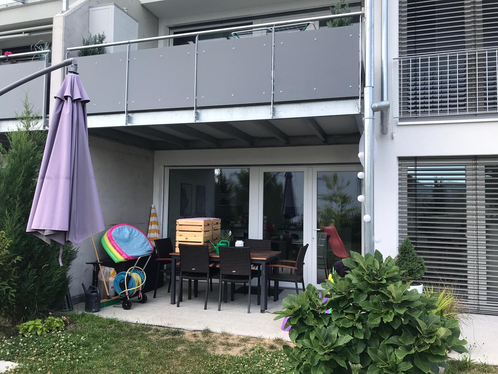 Terrasse einer Wohnung mit Esstisch, Stühlen, lilafarbener Sonnenschirm und Holzkisten