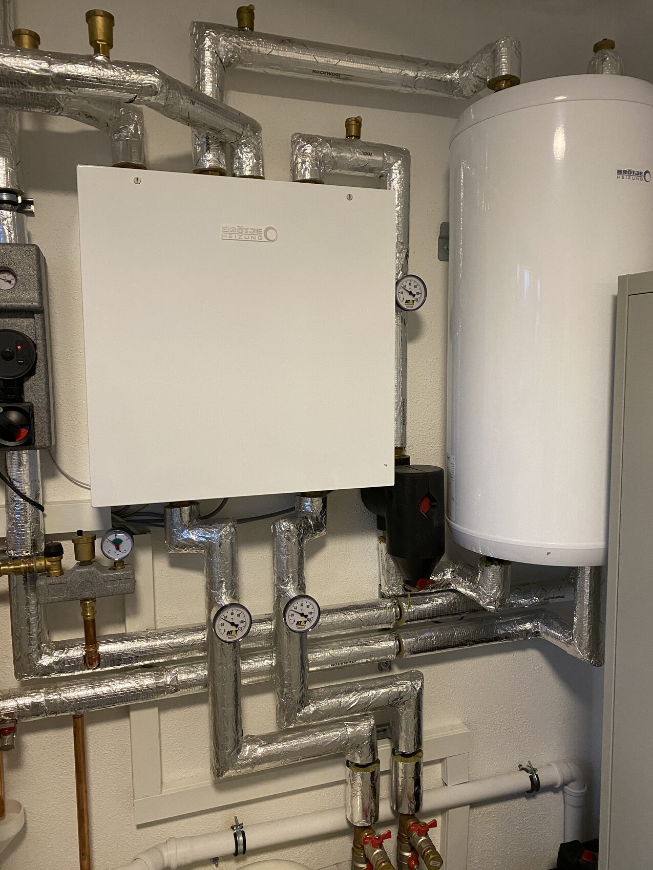 Heizungsraum mit isolierten Rohren, weißem Kessel und Warmwasserspeicher; Messgeräte sichtbar.