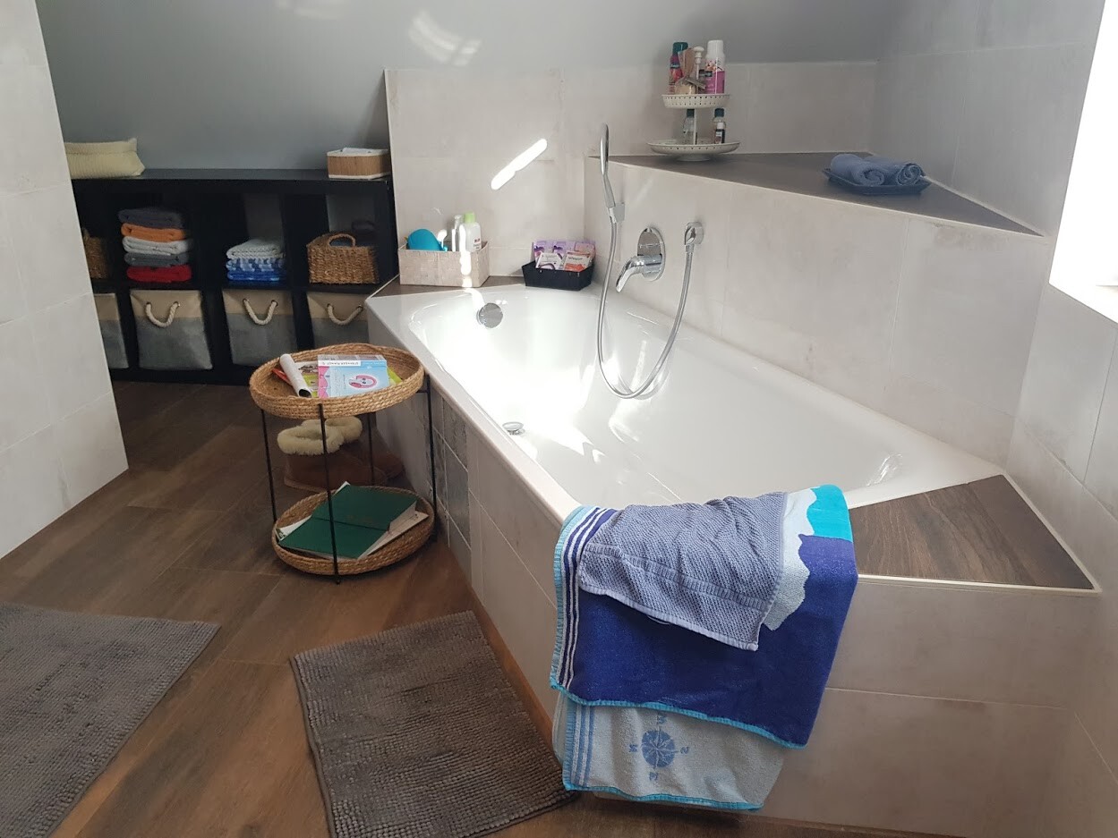 Modernes Badezimmer: weiße Badewanne, Duschbrause, graue Fliesen, Körbe und Handtücher.