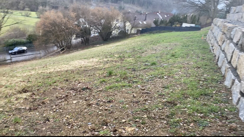 Hangfläche mit Gras, Kies und Steinmauer rechts; Straße, Auto und Häuser im Hintergrund.