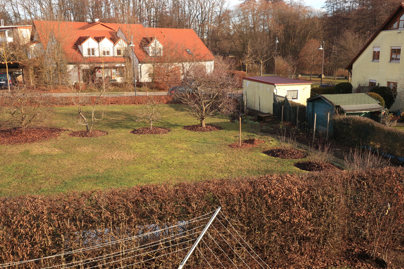 Garten mit Bäumen, Rasenfläche und Häusern mit roten Dächern im Hintergrund.