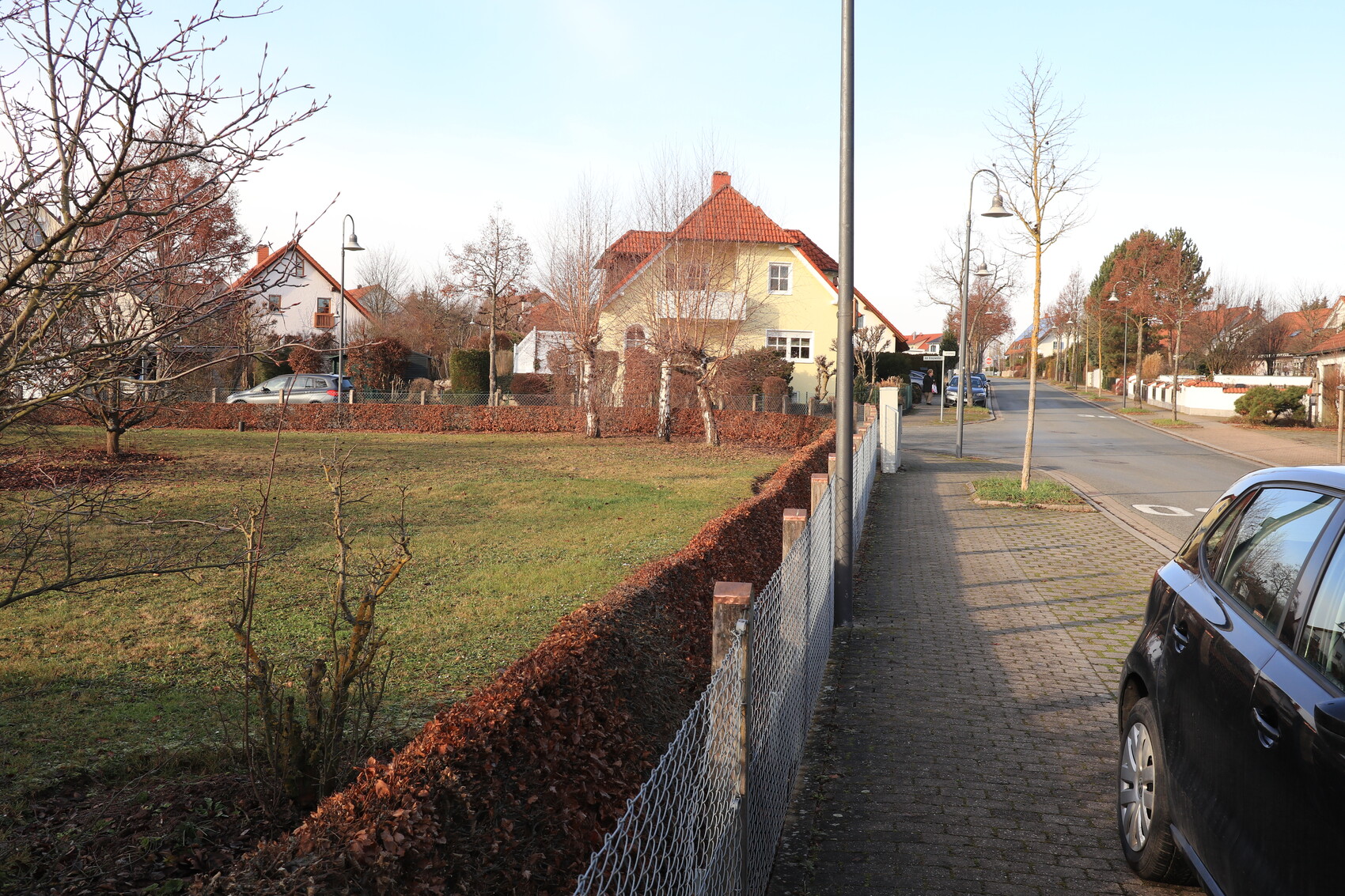 Vorstadtstraße mit gelbem Haus, Gehweg rechts, braune Hecke, Laternen und parkendem Auto.