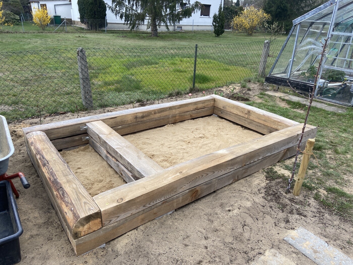 Großer Holz-Sandkasten im Garten, Sand innen, daneben Glas-Gewächshaus