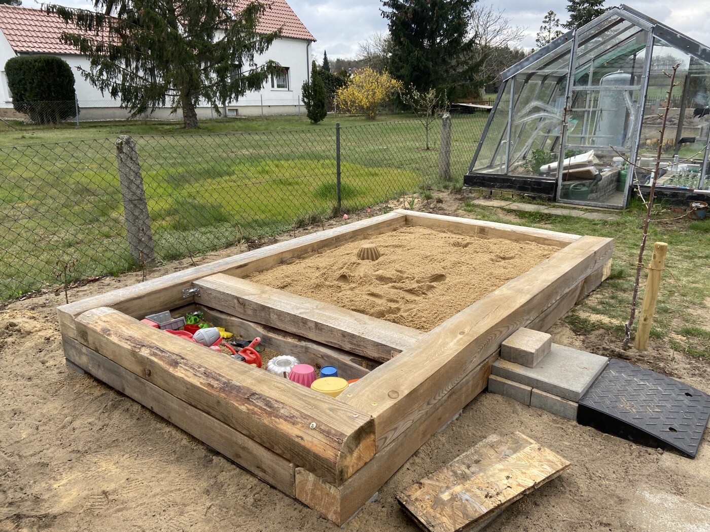 Rechteckiger Holzsandkasten im Garten mit Sand und Spielzeug; Gewächshaus im Hintergrund.