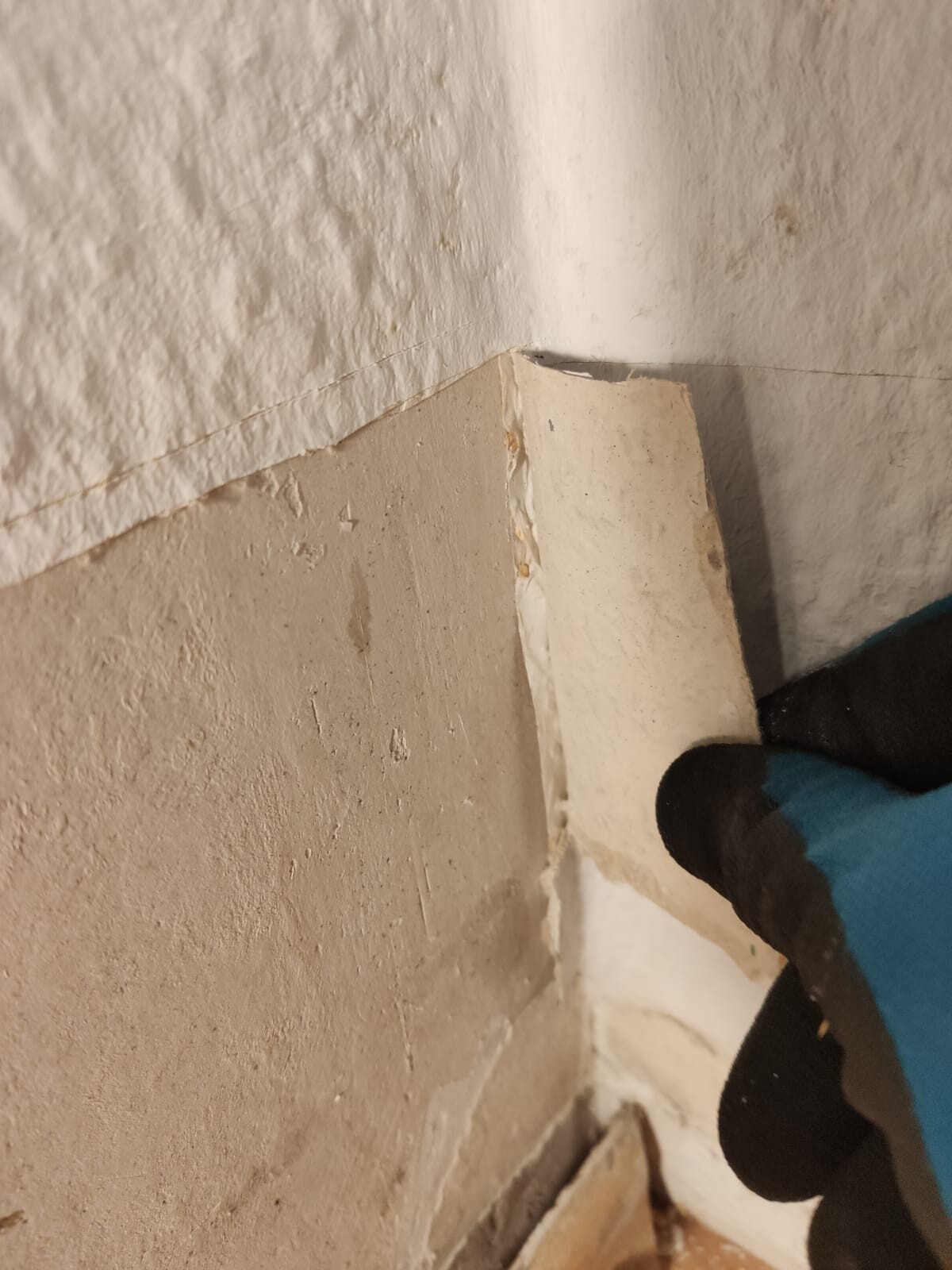 Nahaufnahme einer beschädigten Innenwand-Ecke mit abplatzendem Putz und blauer Handschuh