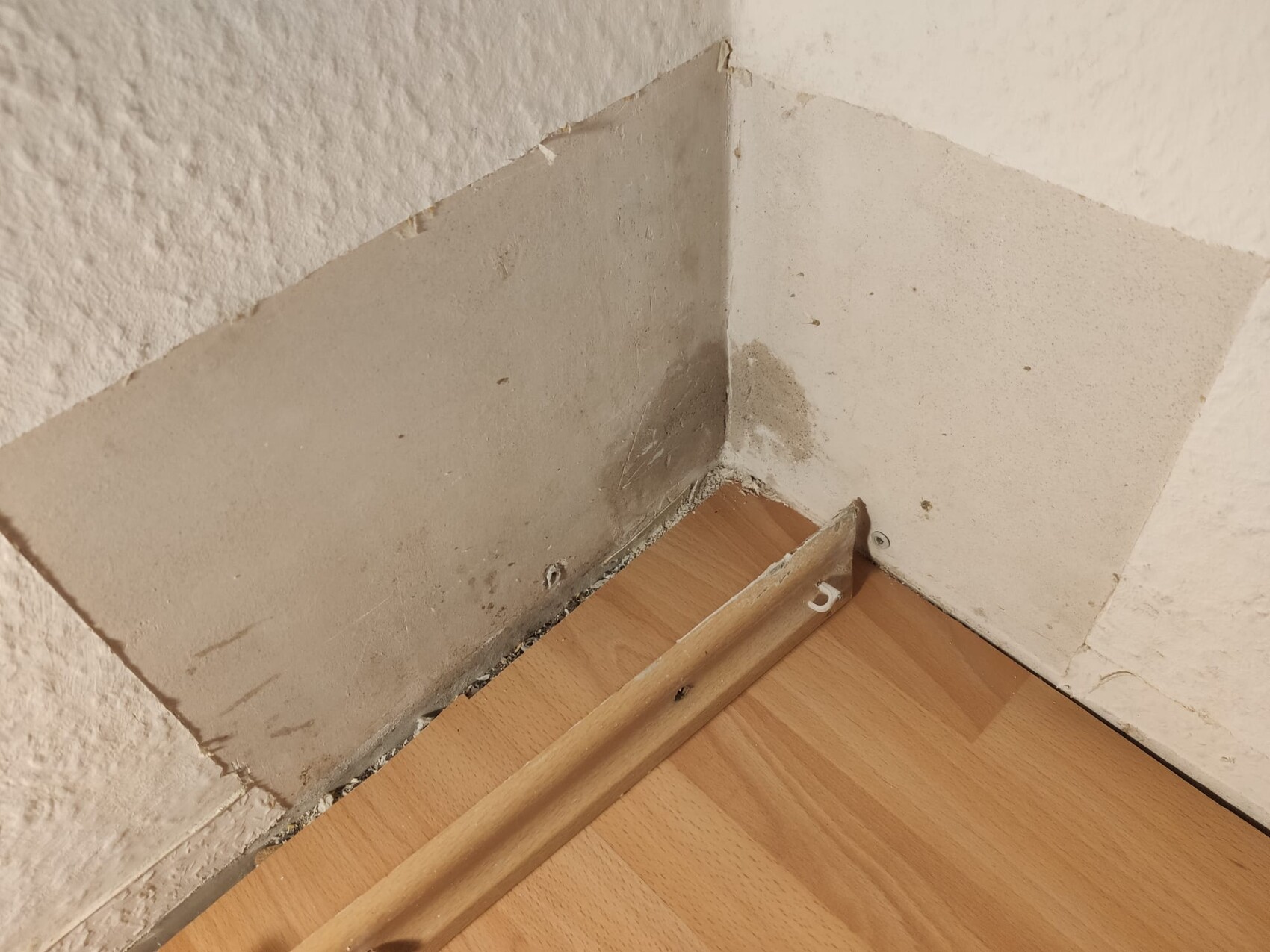 Ecke eines Zimmers mit beschädigter Wand, Putzbrüche und Laminatboden, lose Sockelleiste.