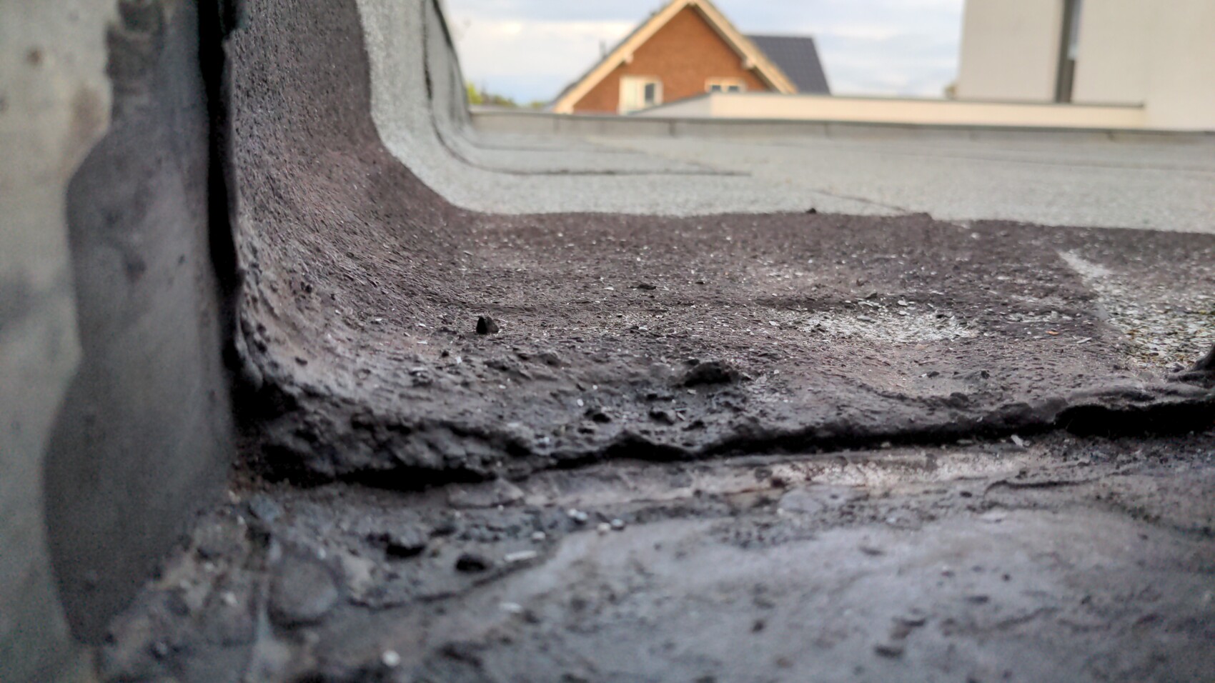 Nahaufnahme einer bitumenbedeckten Dachkante mit Teerresten; Häuser im Hintergrund.