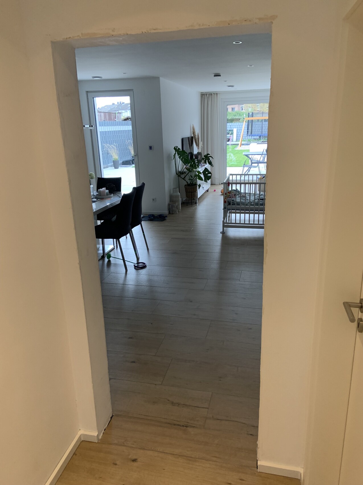 Heller offener Wohnraum mit Esstisch, Stühlen, Pflanze, Babybett und Gartenblick.