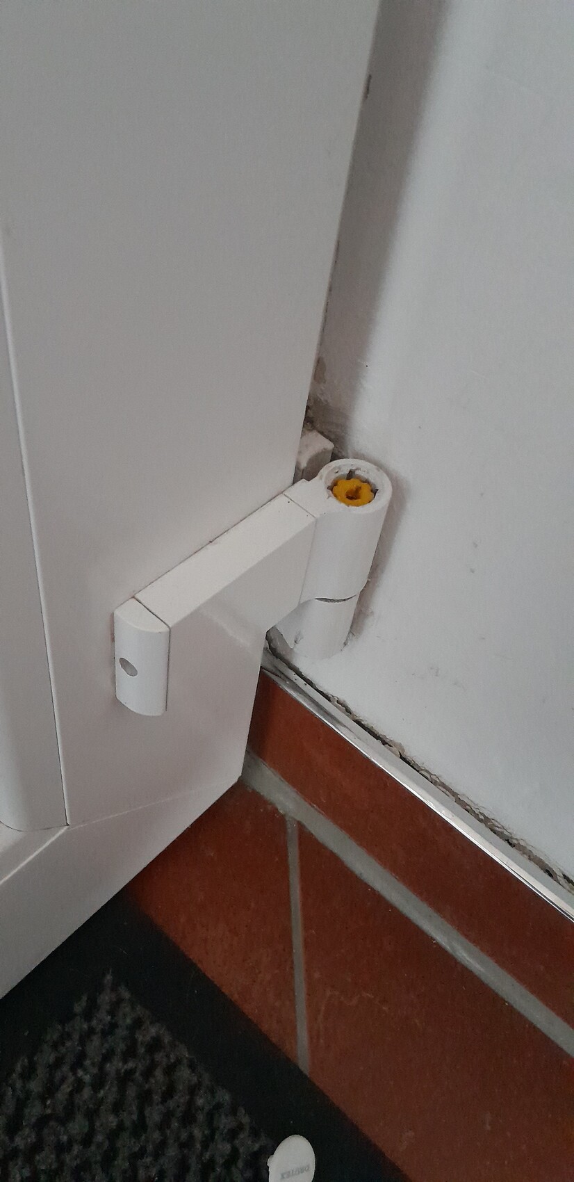 Weißes Tür-Scharnier am unteren Rahmen, gelber Schraubenkopf sichtbar, Fliesenboden.