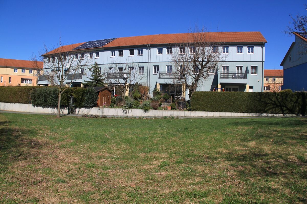 Mehrfamilienhaus mit blauer Fassade, orange Dach, Solarmodule auf dem Dach, Garten davor.
