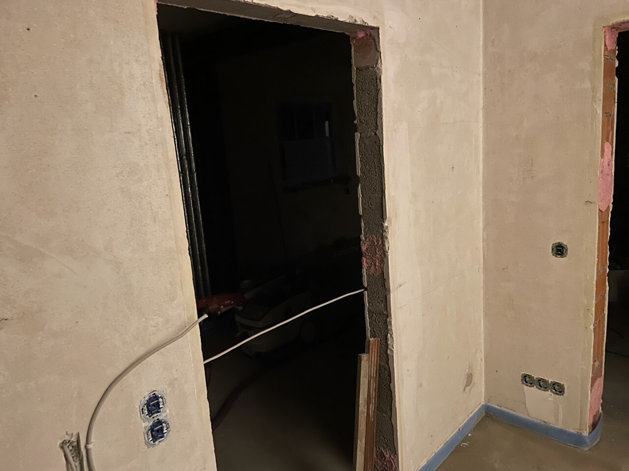 Baustelle: Offene Türöffnung zu dunklem Raum, grob verputzte Wände, Kabel und Steckdosen sichtbar.