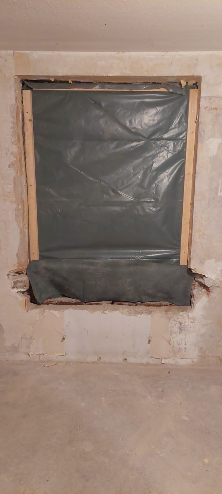 Kellerfenster, von schwarzer Plastikfolie bedeckt, von Holzrahmen eingefasst, in roher Betonwand.