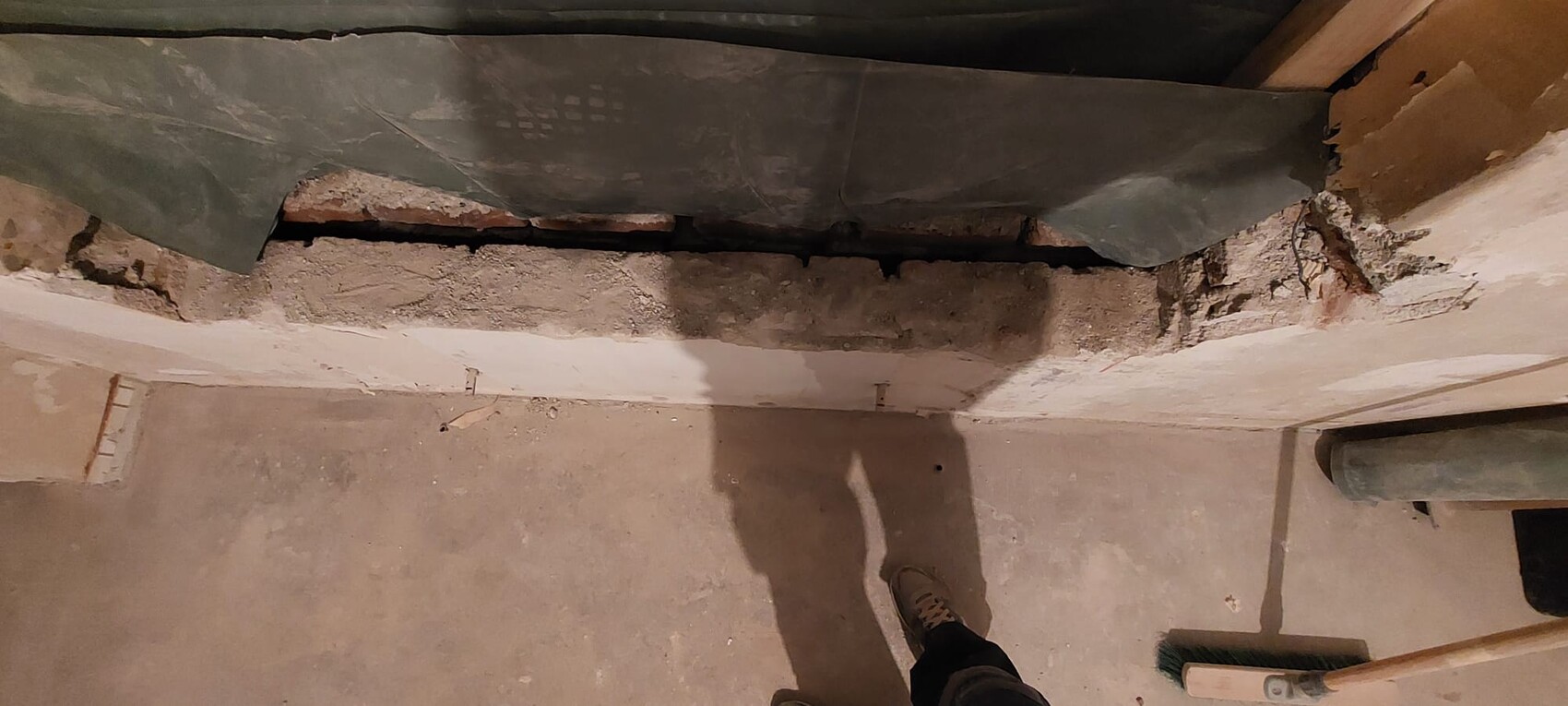 Kellerbaustelle: beschädigte Wand mit Spalt, Putz bricht ab; Besen und Schuh sichtbar.