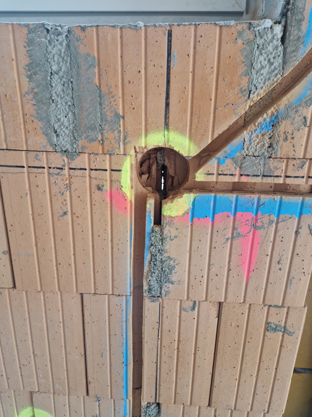 Hohlziegelwand mit zentralem Rundloch, Zementresten und bunten Neon-Sprühmarkierungen.