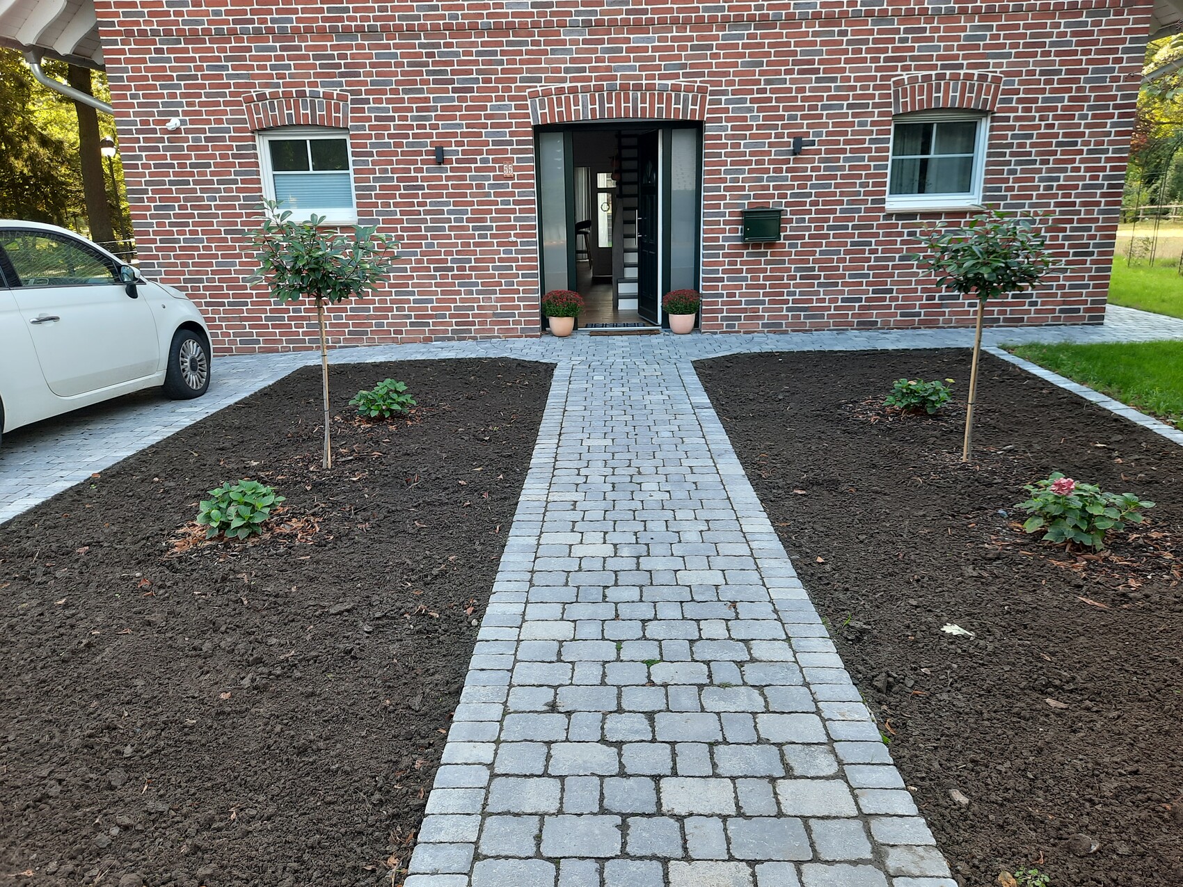 Rotes Backsteinhaus; gepflasterter Weg zur offenen Haustür, Beete mit zwei Bäumen.