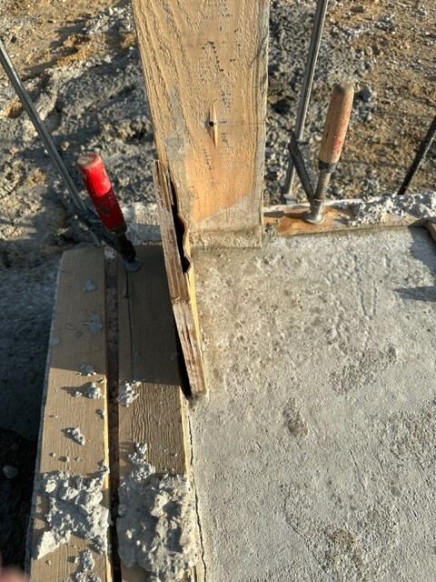 Baustellenszene: Holzform am Betonfundament, Bewehrungsstäbe ragen seitlich, grober Beton.