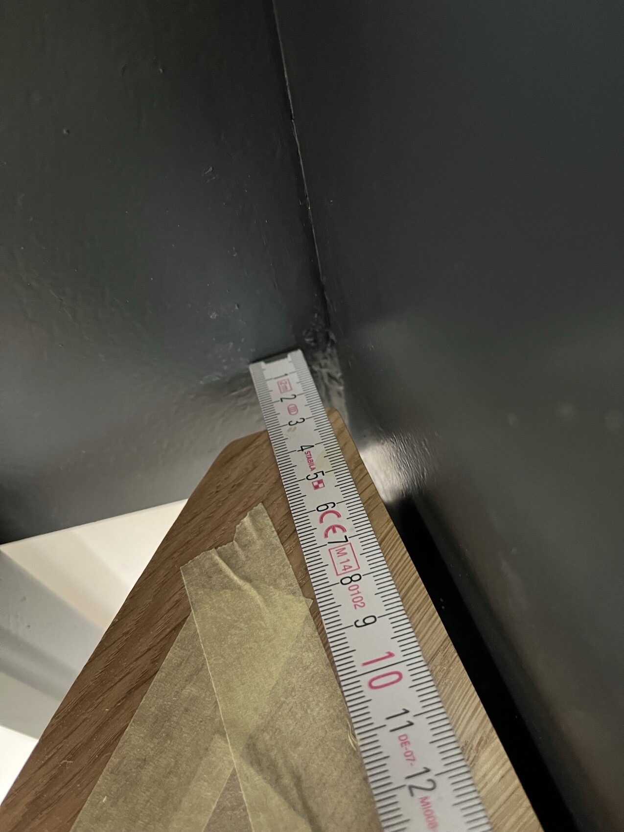Diagonales Maßband an der Ecke zweier dunkler Wände neben einer Holzplatte; Klebeband liegt darauf.