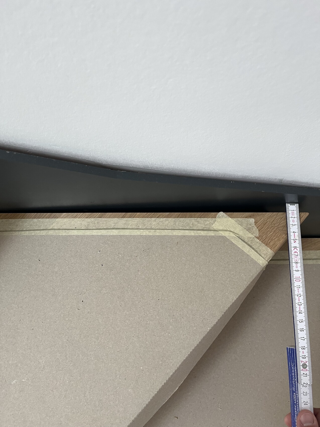 Messband neben Kante einer beigen Spanplatte vor grauer Wand, mit Malerkrepp.