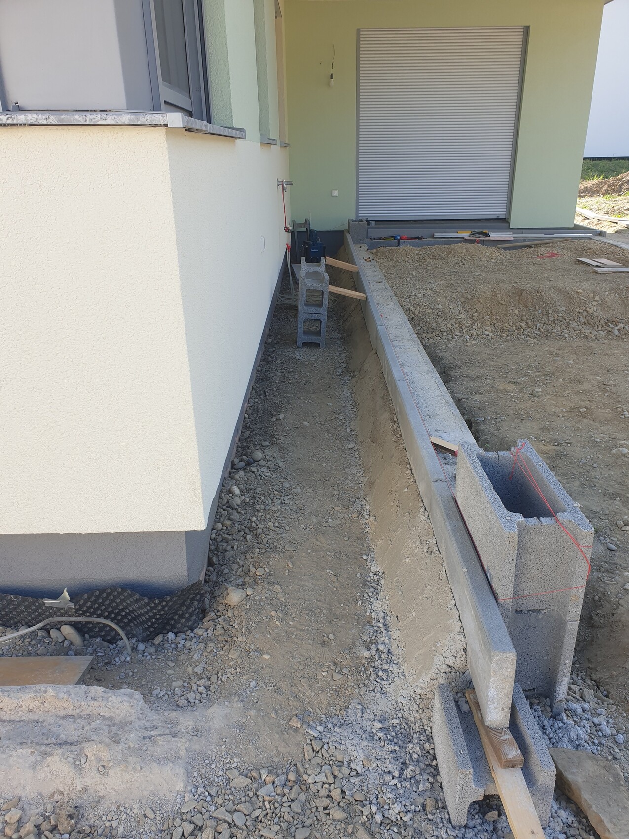 Baugrube neben der Hauswand mit Betonblöcken, Holzlatten und Werkzeugen auf der Baustelle.