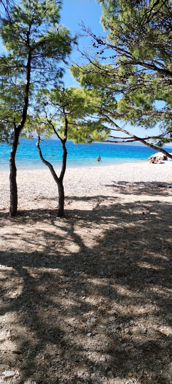 Kiesstrand mit Schatten von Kiefern, türkisblaues Meer, einige Badegäste.