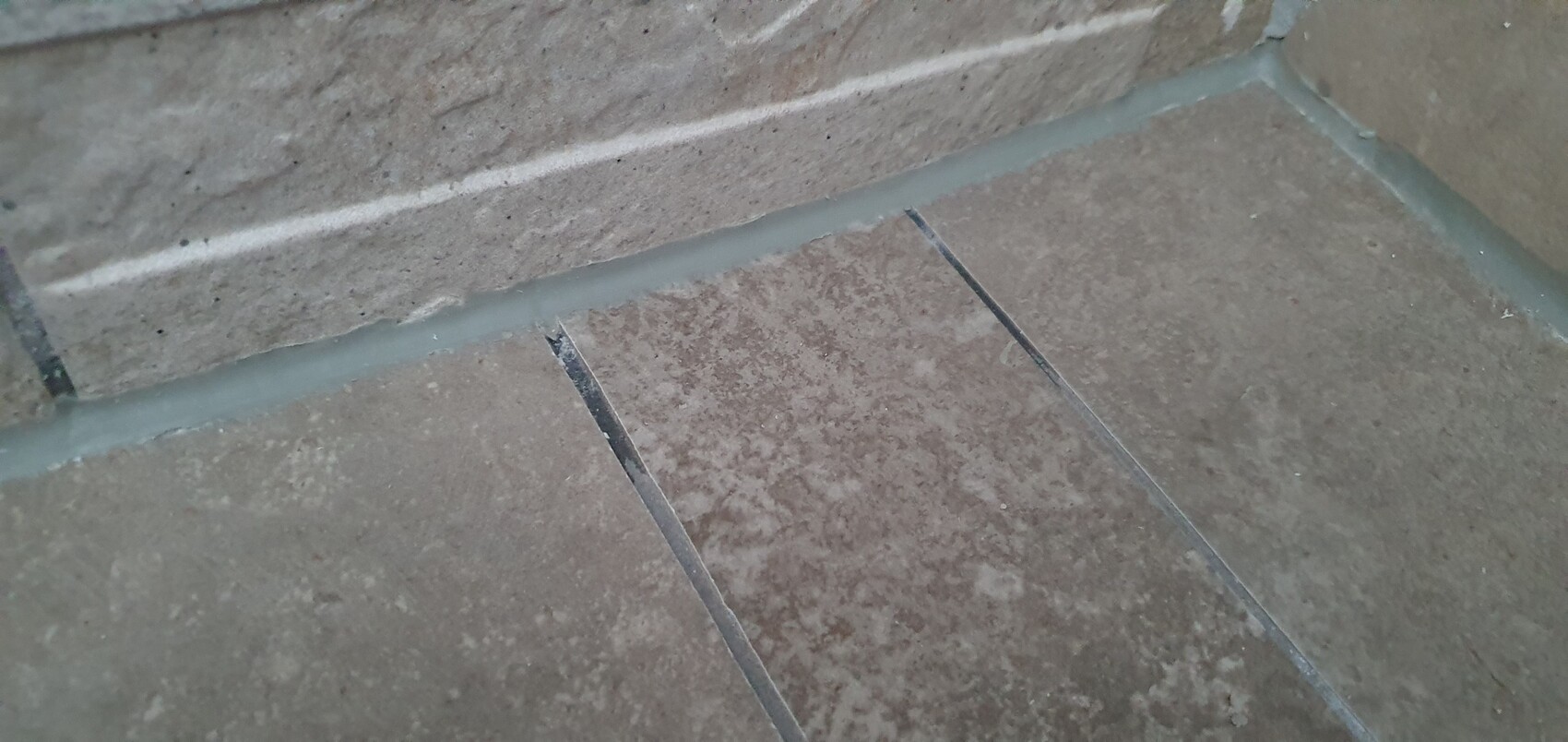 Braune Bodenfliesen mit grauen Fugen; Ecke einer Betonwand mit blauem Silikonfugenstreifen.