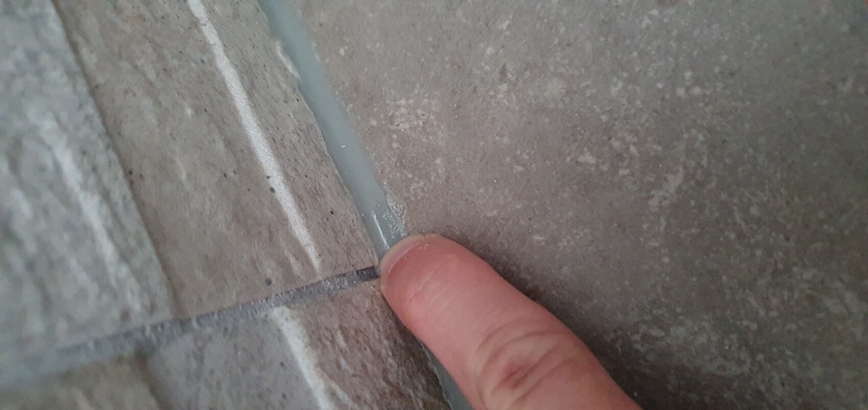 Nahaufnahme eines Fingers, der eine graue Silikonfuge zwischen Fliesen berührt.