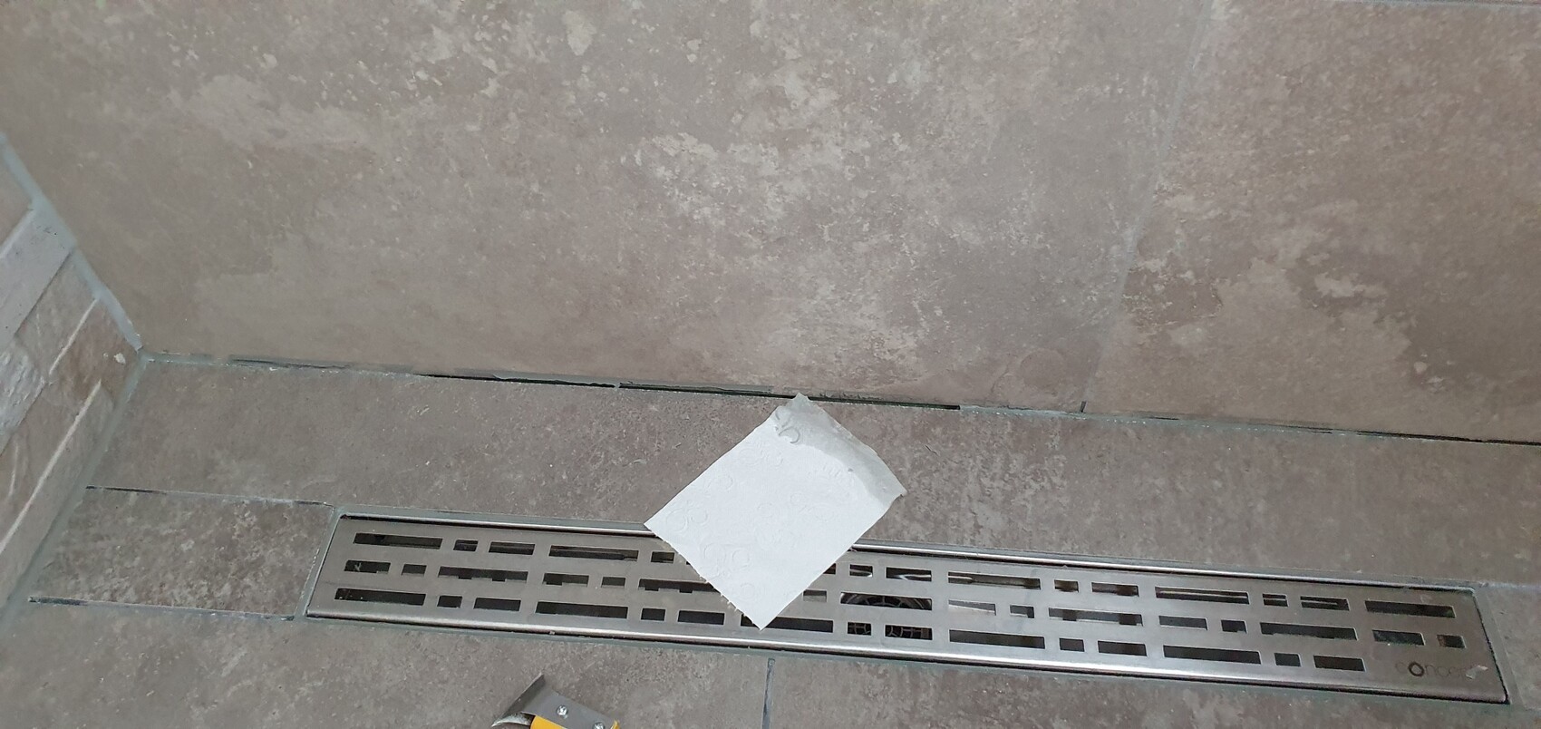 Brauner Fliesenboden mit langem Edelstahl-Drain; ein weißes Papiertuch liegt darauf.
