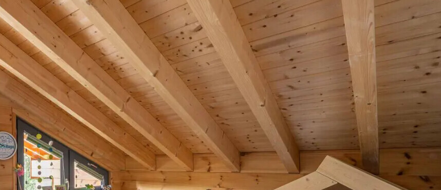 Holzdecke mit sichtbaren Balken, Dachboden-Innenraum; linke Wand mit Poster-Infotafel.