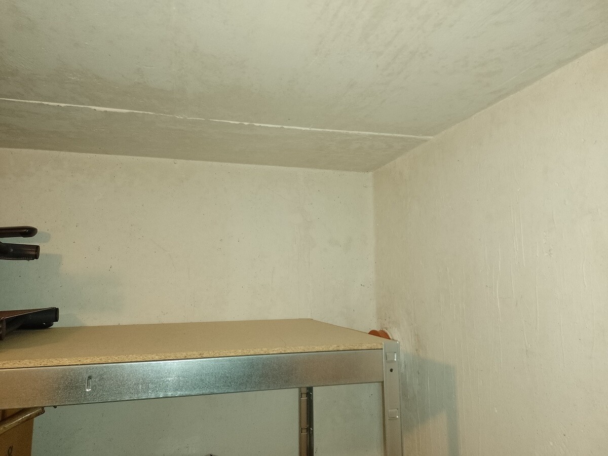 Kellerabstellraum mit Betonwänden und Decke; unten ein Metallregal mit Gegenständen darauf.