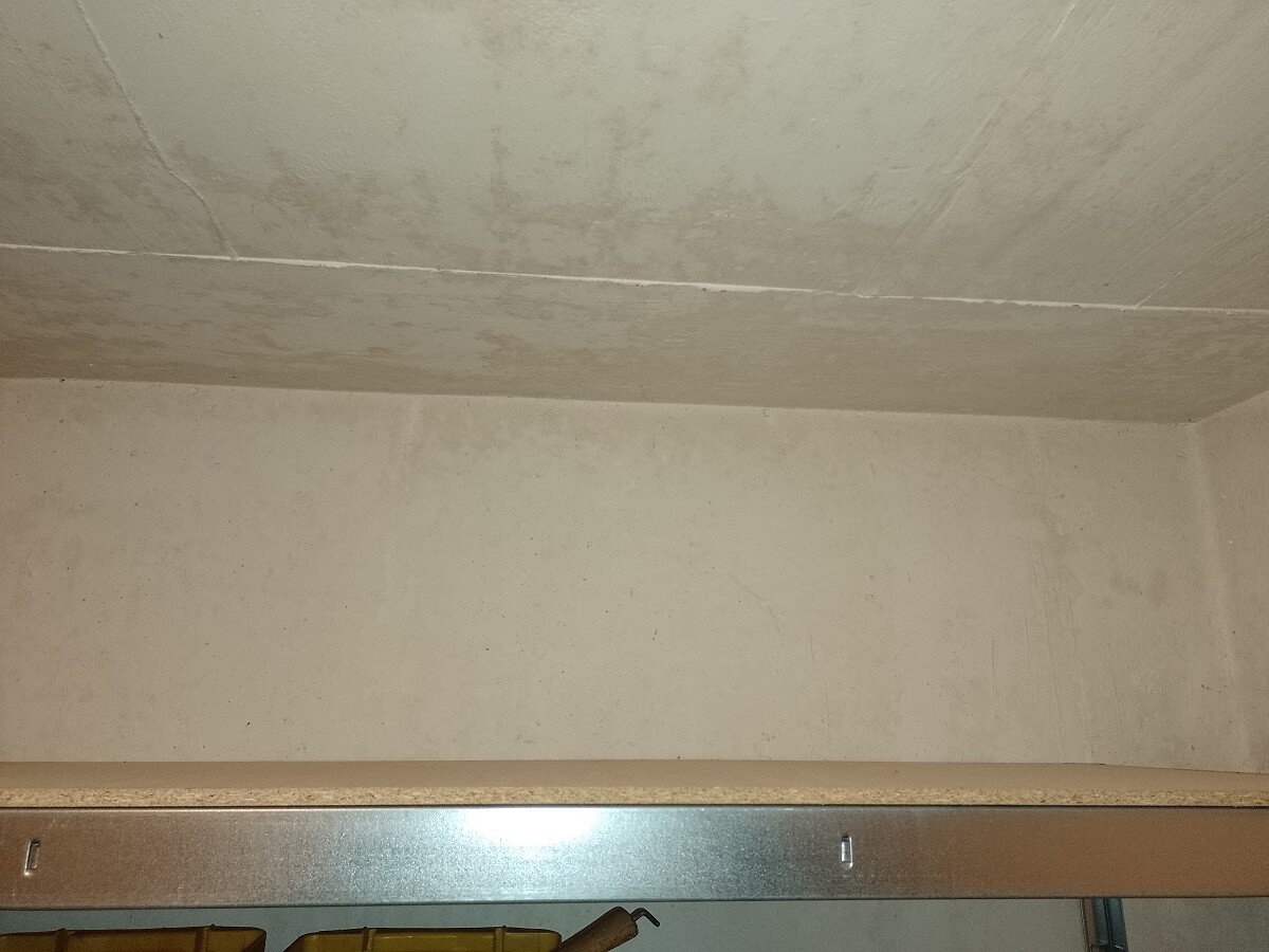 Leerer Kellerraum mit grauer Betonwand und Decke, Metallregal unten und gelben Kästen.