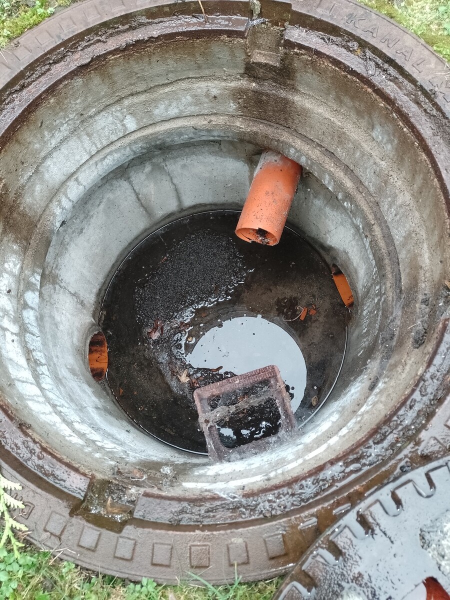 Offener Schacht mit schwarzem Wasser und Schlamm; ein orangefarbenes Rohr ragt hinein.