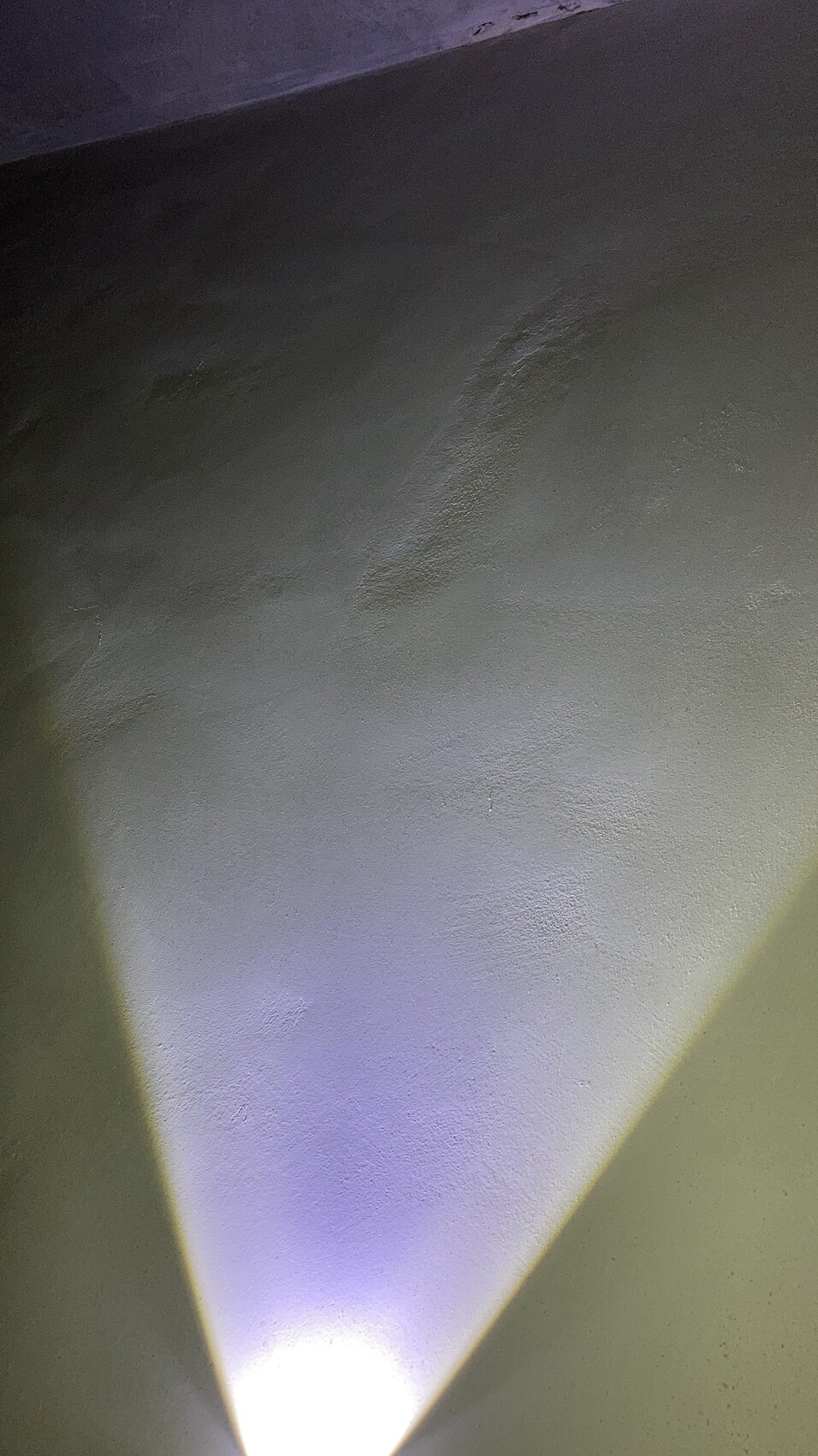 Rauhe grüne Wand mit einem von unten aufhellenden farbigen Lichtkegel in V-Form.