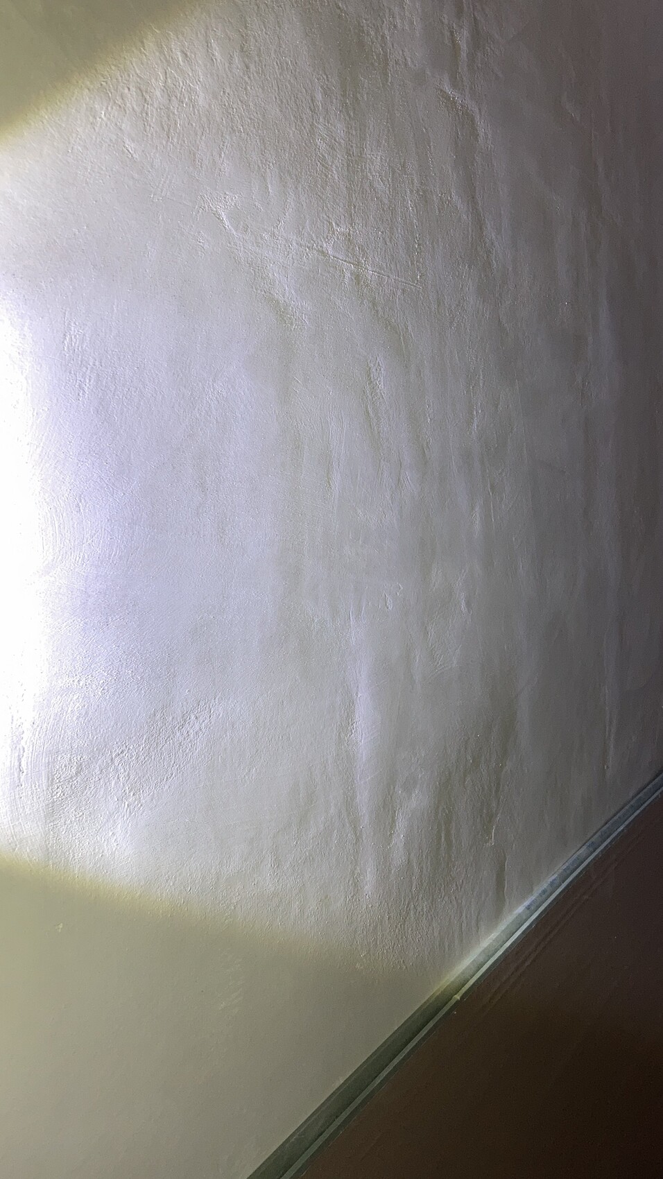 Weiße Wand mit körniger Putzstruktur; unten Glas-/Metallgeländer am dunklen Boden.