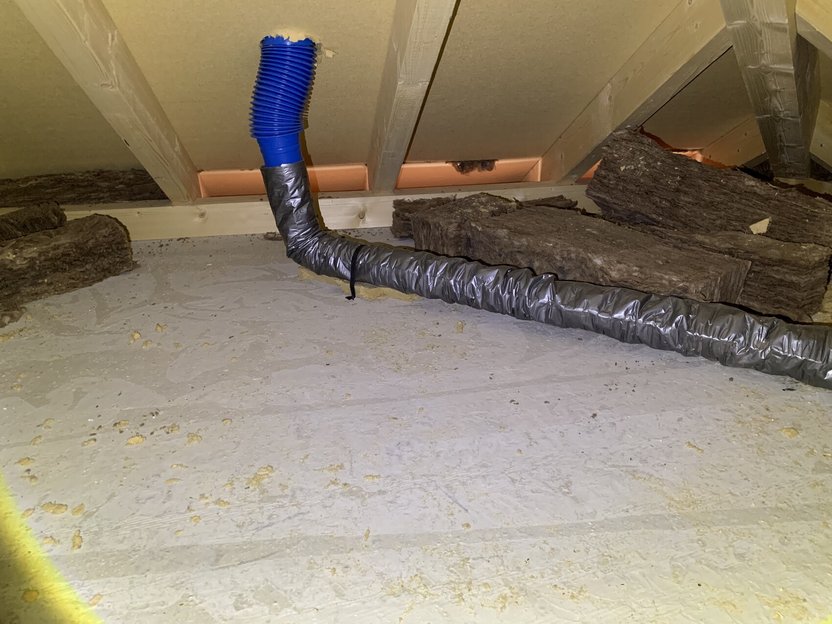 Dachboden: blauer flexibler Schlauch verbindet sich mit silberner Isolierung, Holzbalken sichtbar.