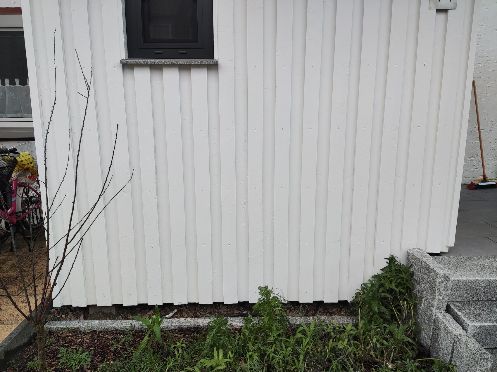 Weiße Wand aus vertikalen Holzpaneele, kleines Fenster, Gartenpflanzen und Besen davor.