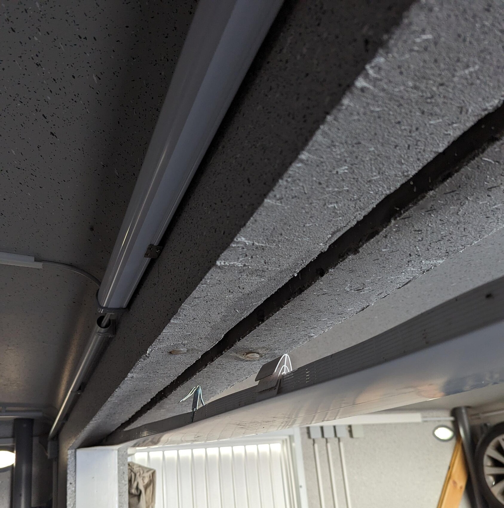 Unterseite einer rauen Beton-Decke in einer Werkstatt, mit Leuchtstoffröhre und Kabelclips.