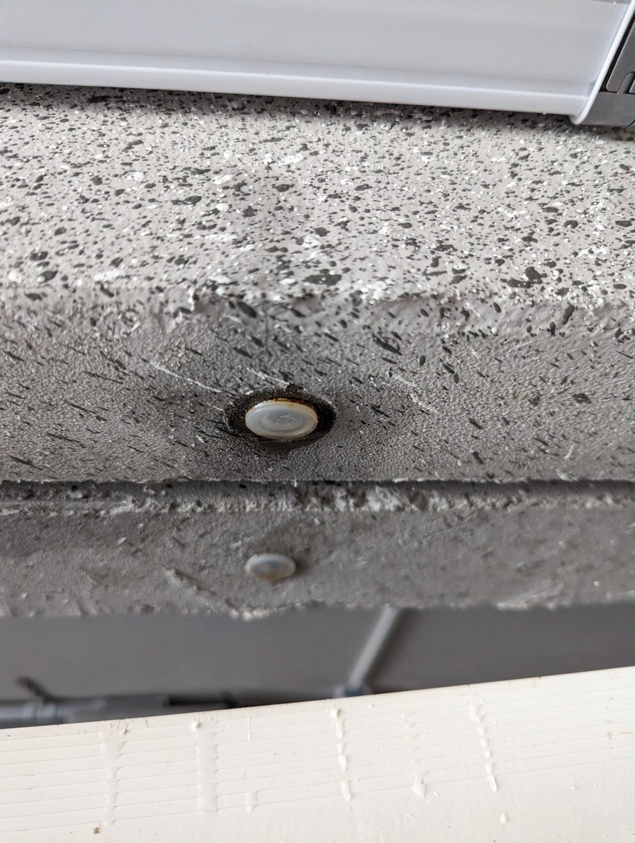 Runde weiße Abdeckung in dunkler Ringöffnung auf rauer grauer Oberfläche; daneben kleiner Knopf.