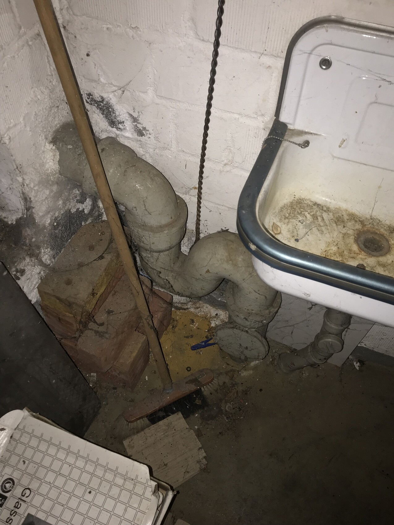 Kellerbad/Werkstattraum: schmutzige Spüle, große graue Abflussrohre und Unrat am Boden.