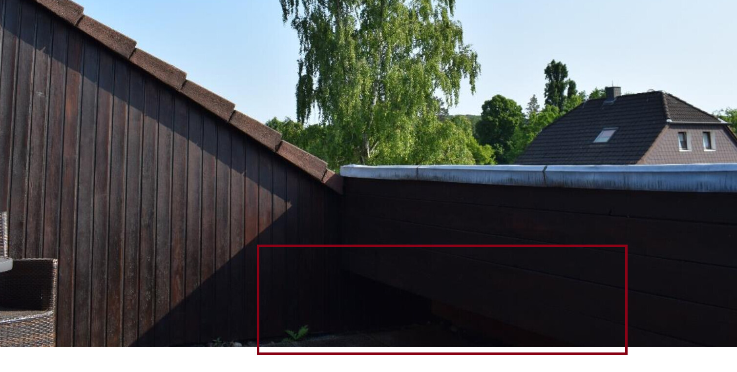 Dunkle Holzverkleidung an einer Dachkante, Schatten unten; Bäume und Haus im Hintergrund.