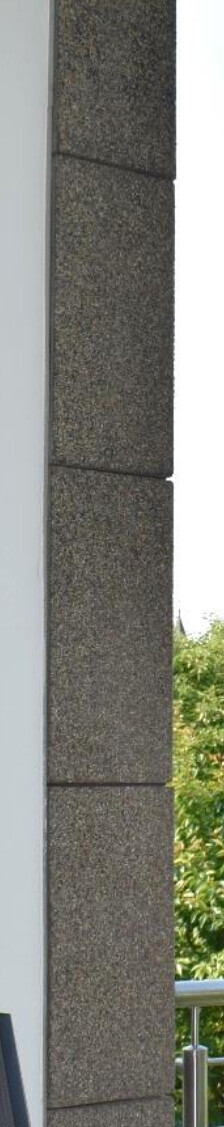 Außenwand-Ecke: graue Steinblöcke neben heller Wand, Geländer und grüne Bäume dahinter.