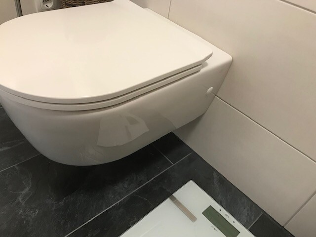 Moderne weiße wand-Toilette mit geschlossenem Deckel; digitale Waage auf dunklem Fliesenboden.
