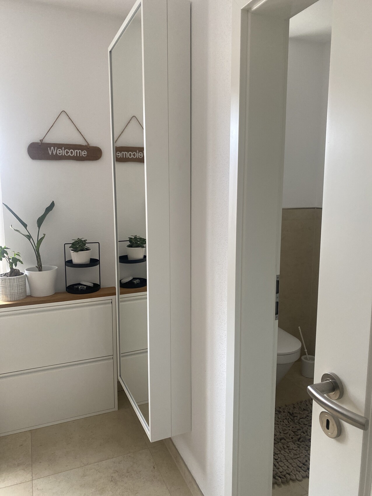 Flur mit weißem Schrank mit Spiegel, Willkommensschild, Pflanzen; Badezimmer mit Toilette sichtbar.