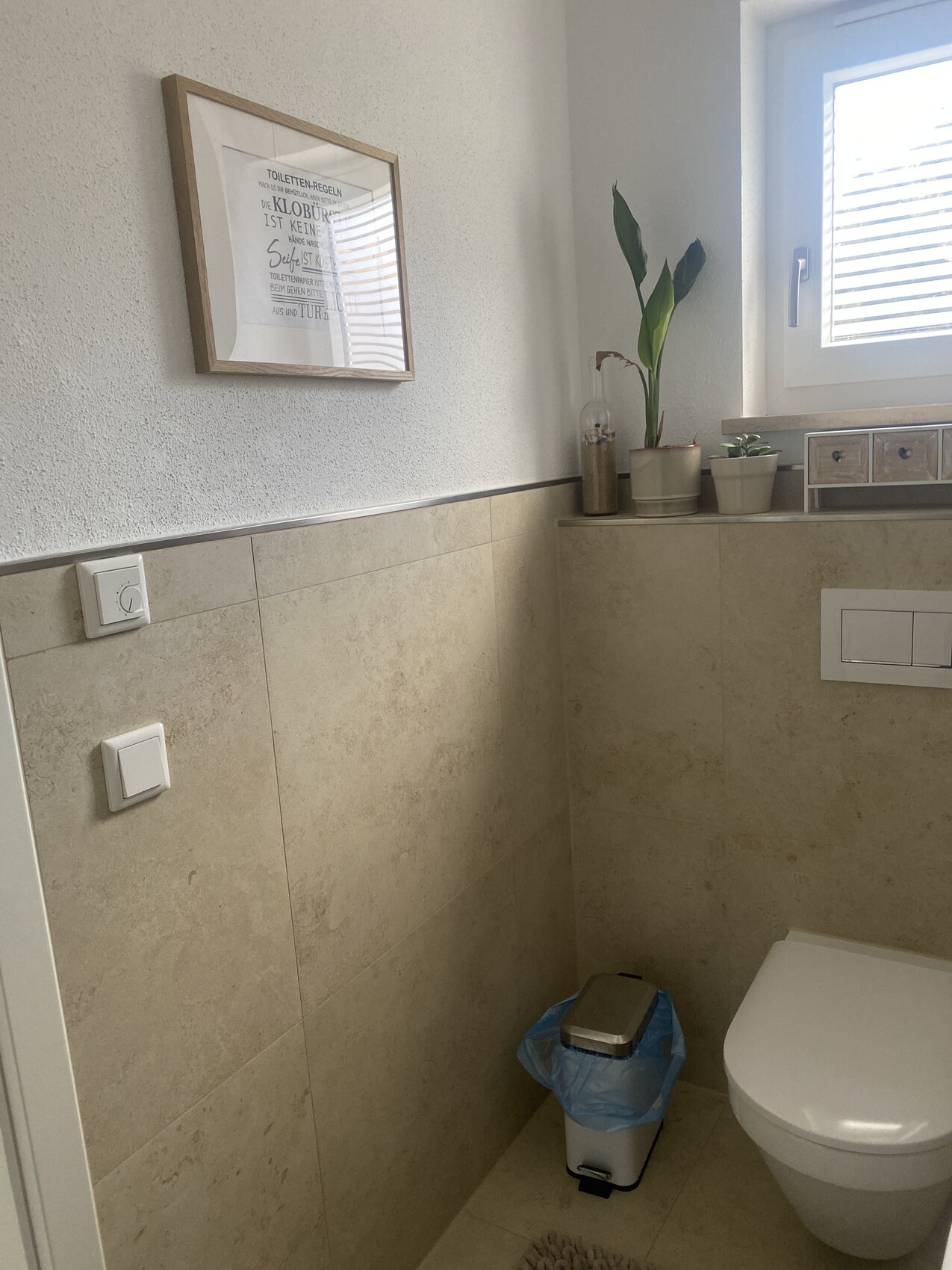 Badezimmer: Beige Fliesen, Wandbild, Fenster mit Pflanze, Toilette, Mülleimer.
