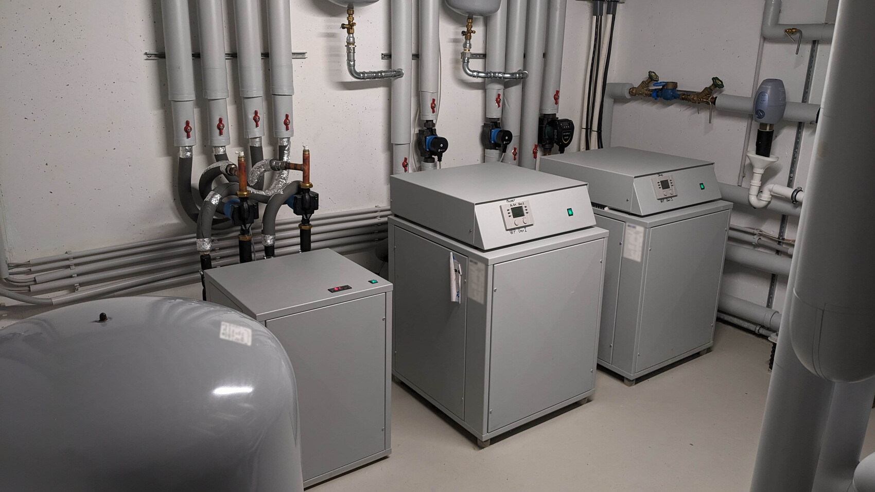 Drei industrielle Waschmaschinen in einem Technikraum mit Rohren, Ventilen und Pumpen